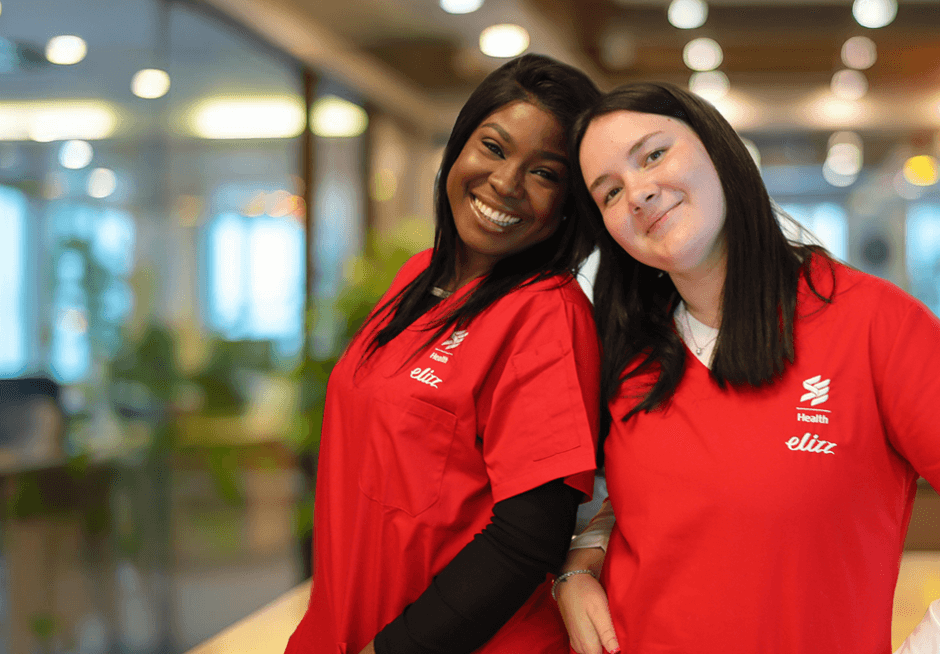 A healthcare worker in red scrubs stands outside a home with arms crossed, smiling at the camera.