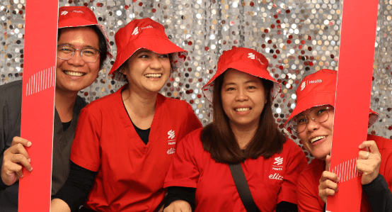Four healthcare workers in red scrubs and hats smile together while posing with red photo-frame props in front of a festive backdrop.